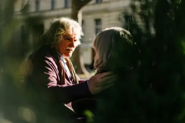 A man with white, curly hair and a mustache gently places his hand on another person's shoulder, offering a comforting response. This scene illustrates react vs respond examples.