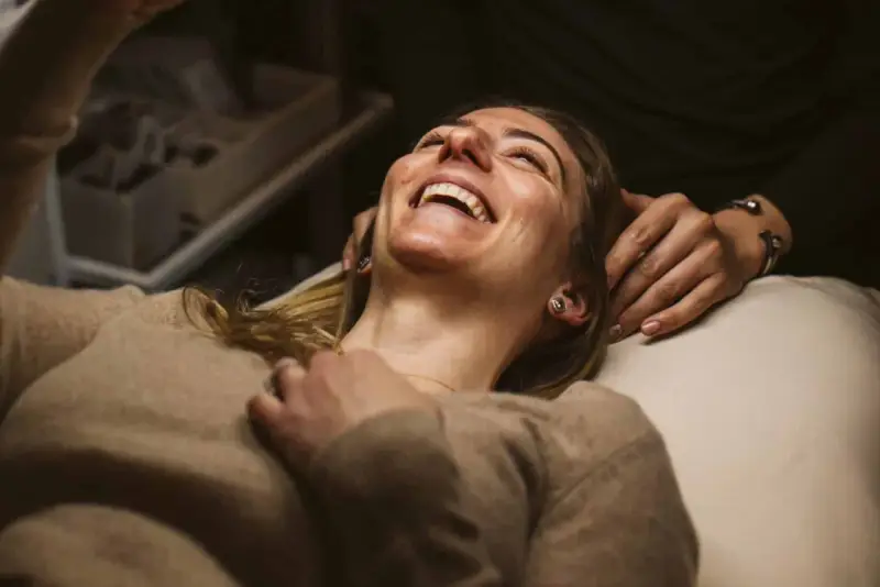 A woman laughs joyfully during a holistic facial treatment, her head tilted back as hands gently support her.